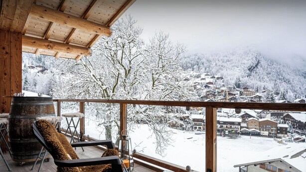 A wooden deck with a chair and a table, overlooking a snowy village. - Le Grand Capricia - mountain stay with sauna - OVO Network (La Clusaz)