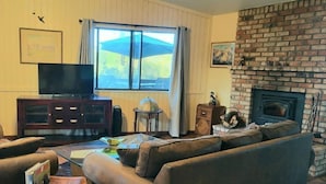 A living room with a brick fireplace, a large window with curtains, a television, and a sofa. - The Getaway (Placerville)