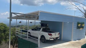 Un camionnette blanche est garée sous une structure bleue dotée d’un toit en métal, entourée de verdure et sous un ciel dégagé.