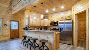 A wooden cabin kitchen with a central island, bar stools, stainless steel appliances, and a large window. - Newly built Luxury cabin with a Game Room, Private Indoor Pool, Hot Tub, Theater (Gatlinburg)