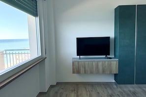A room with a TV on a wooden shelf, a window with a view of the sea, and teal-colored cabinets.