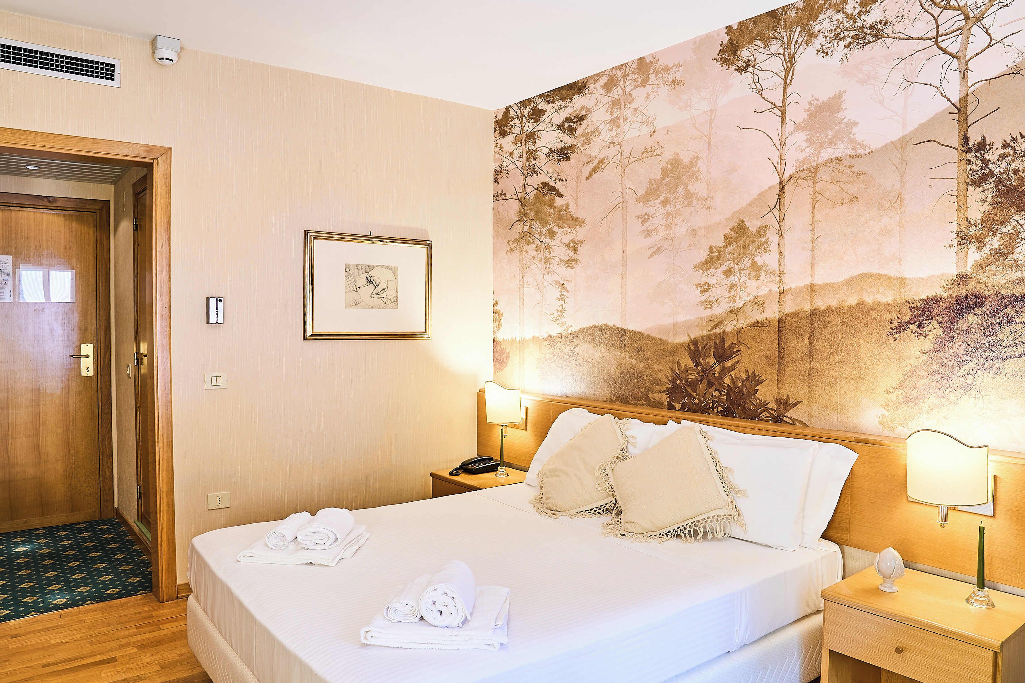 Una habitación de hotel con cama, mesita de noche, lámpara de pared, cuadro enmarcado y una puerta.