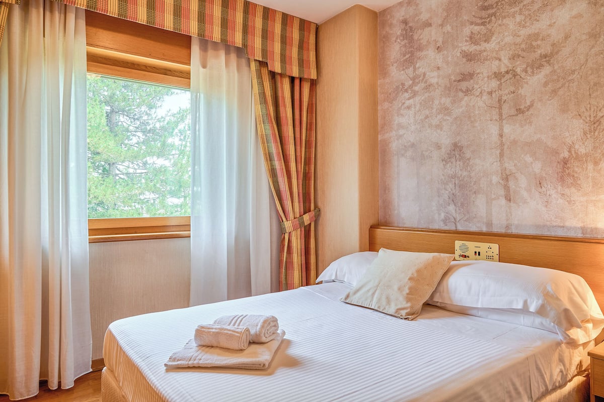 A neatly made bed with white linens and a set of folded towels, a wooden headboard, and a window with sheer curtains and a striped valance.