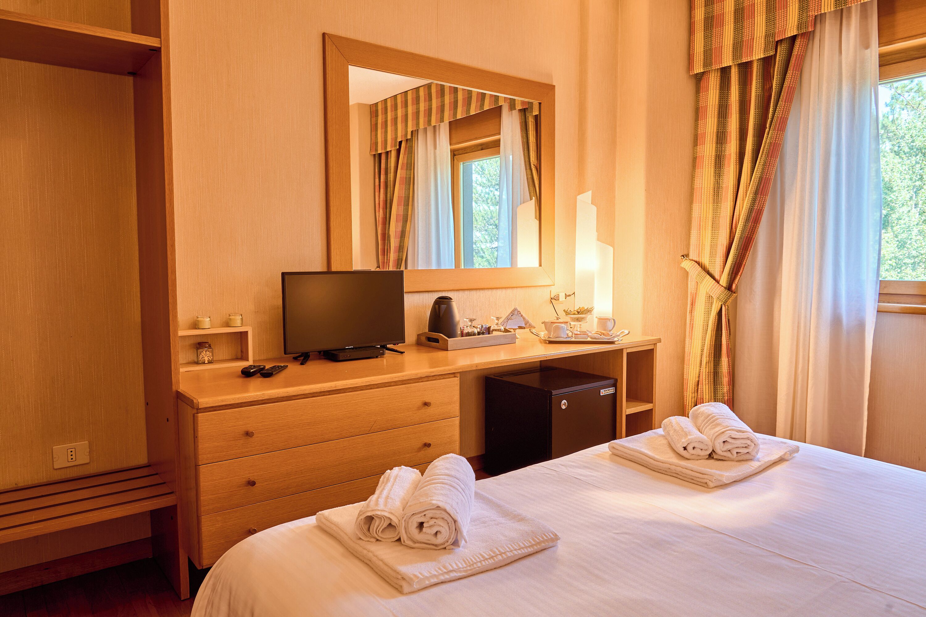 A hotel room with a wooden desk, a television, a bed with towels, a mirror, and a window with curtains.