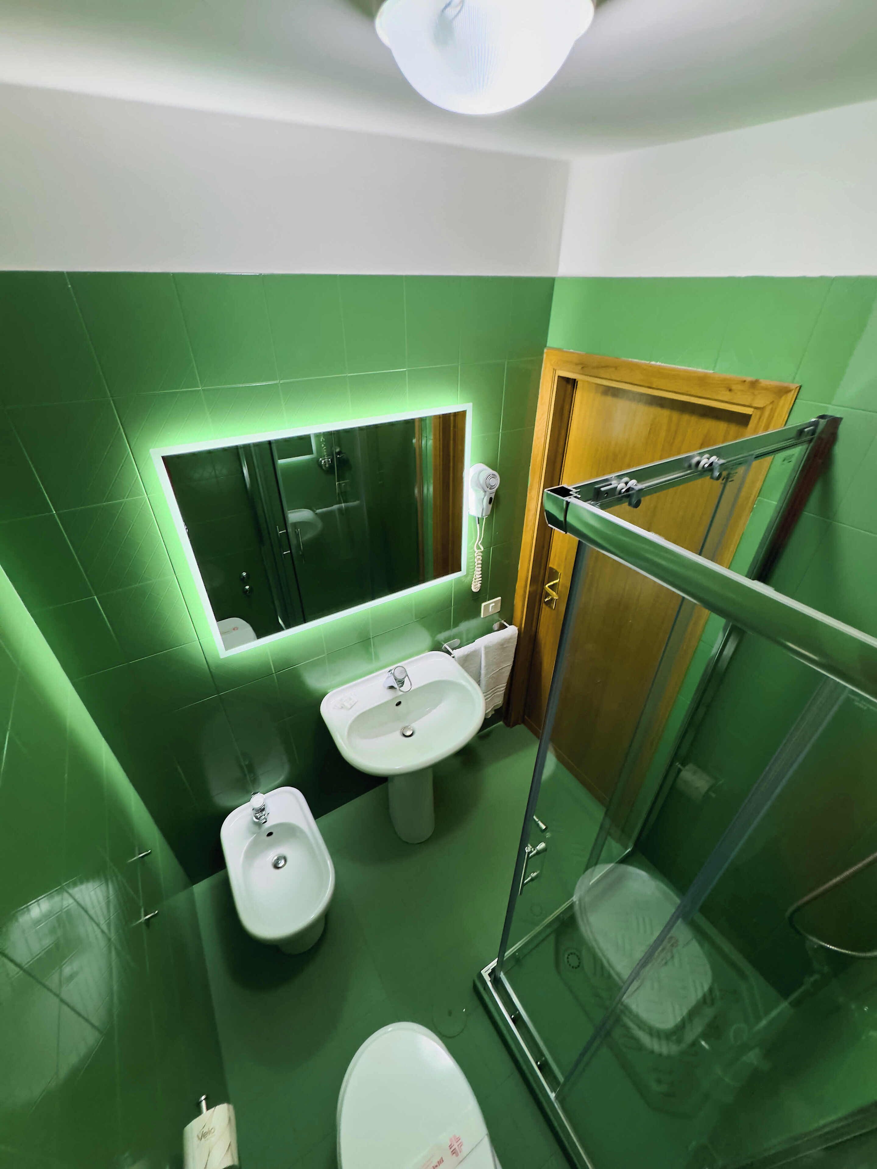 Un baño con paredes verdes, un lavamanos blanco, un bidé, una cabina de ducha de vidrio y una puerta de madera.