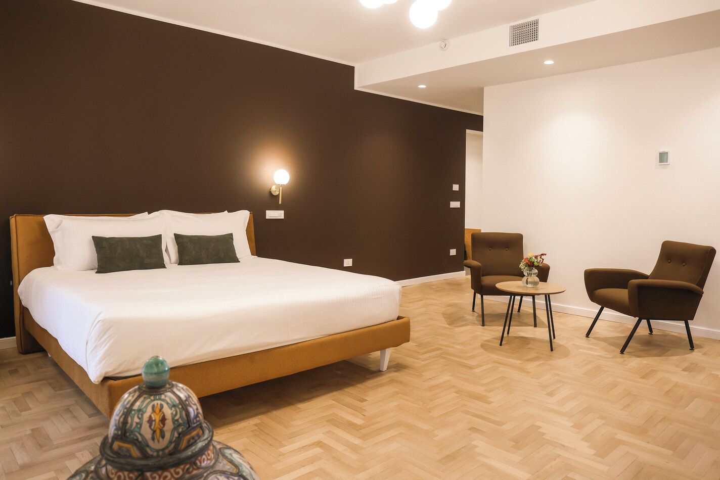Una habitación de hotel moderna con una cama grande, dos sillones, una mesita pequeña y una lámpara decorativa.