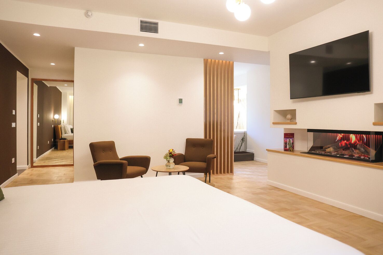 Ein modernes Hotelzimmer mit einem großen Bett, zwei Sesseln, einem kleinen Tisch mit einer Vase, einem flachbildfernseher an der Wand und einem Kamin.