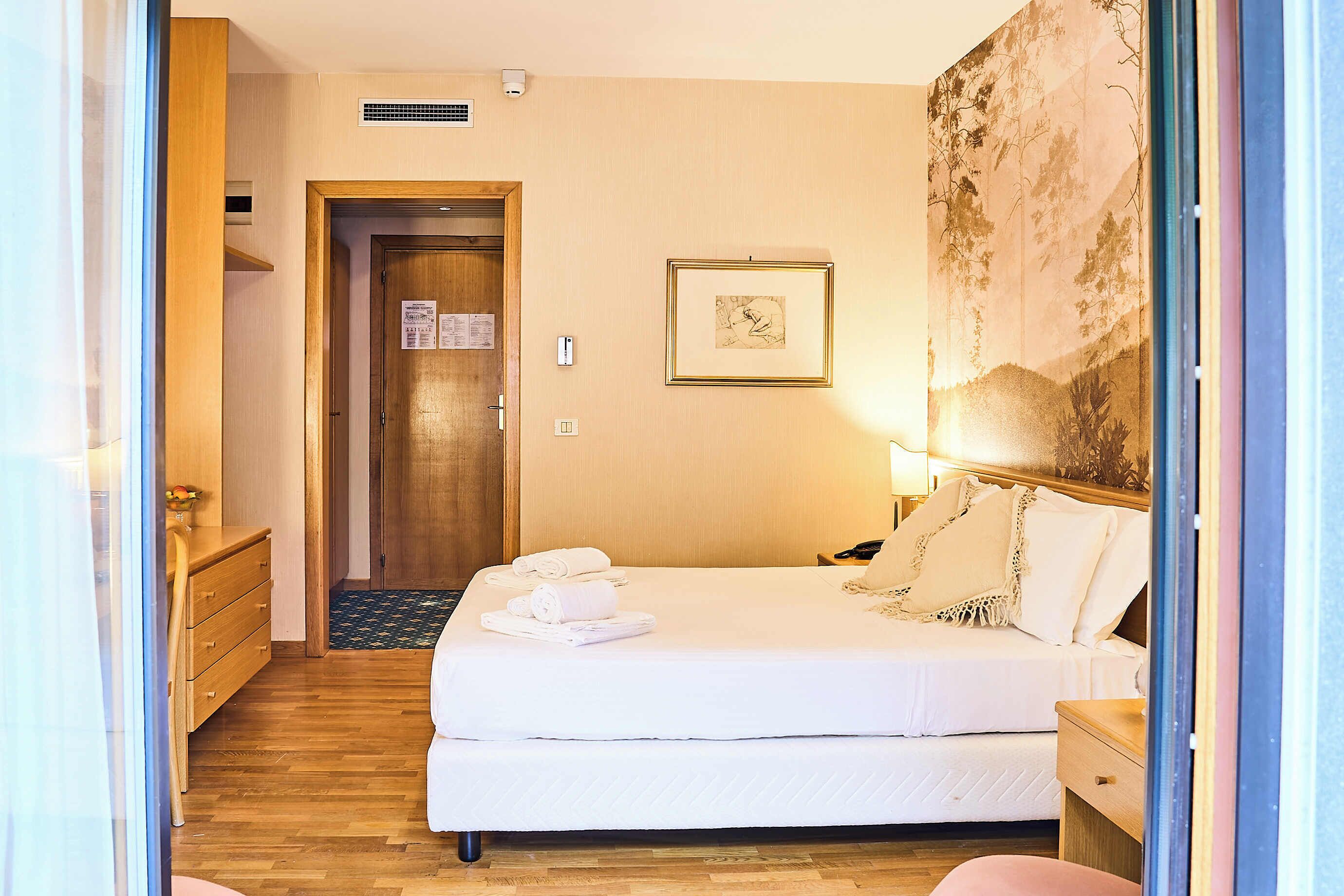 A hotel room with a wooden floor, a bed with white linens, a nightstand, a lamp, a framed picture on the wall, and a door leading to another room.