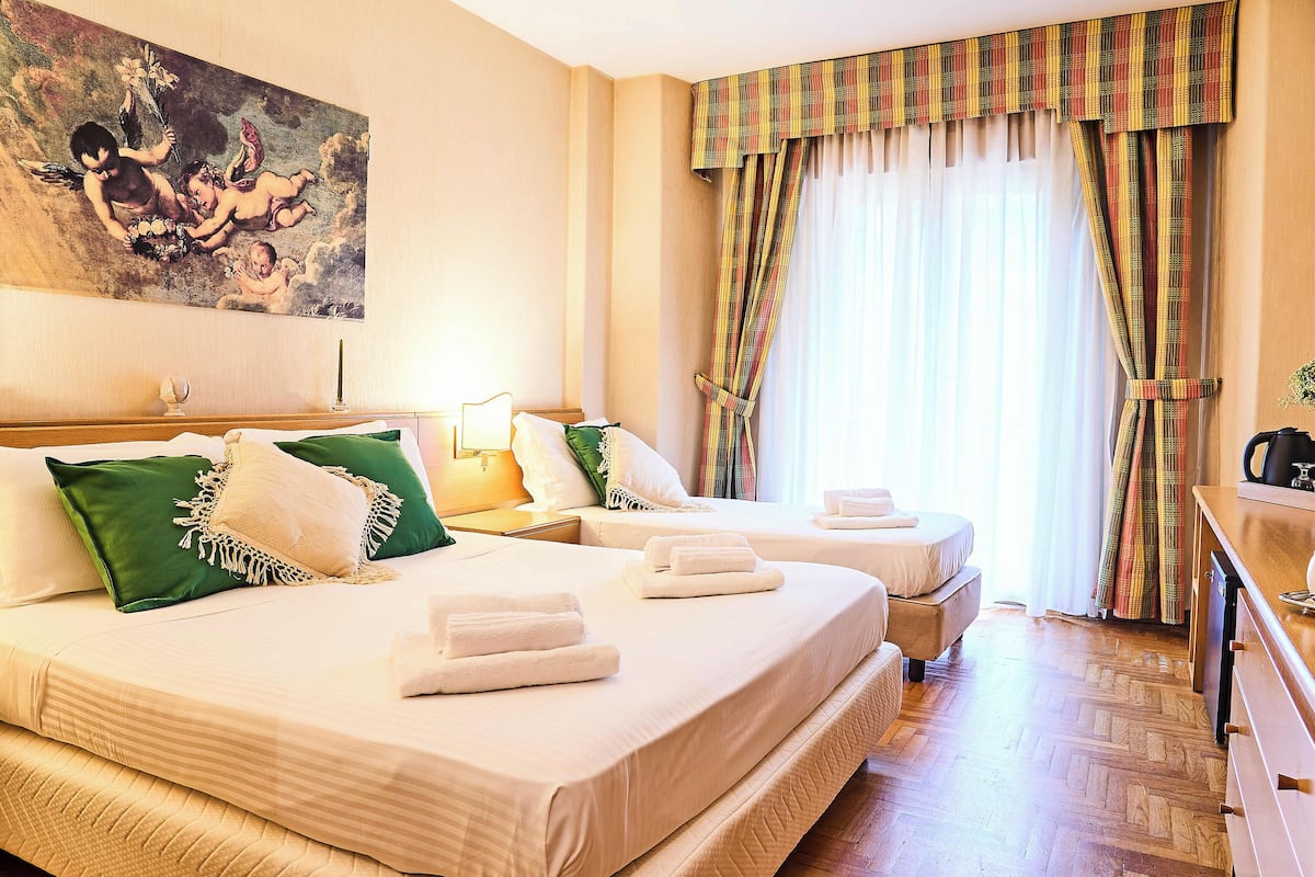 A hotel room with two beds, a painting on the wall, and a window with curtains.