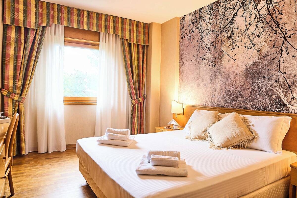 A hotel room with a large bed, two pillows, a nightstand with a lamp, and a window with curtains.