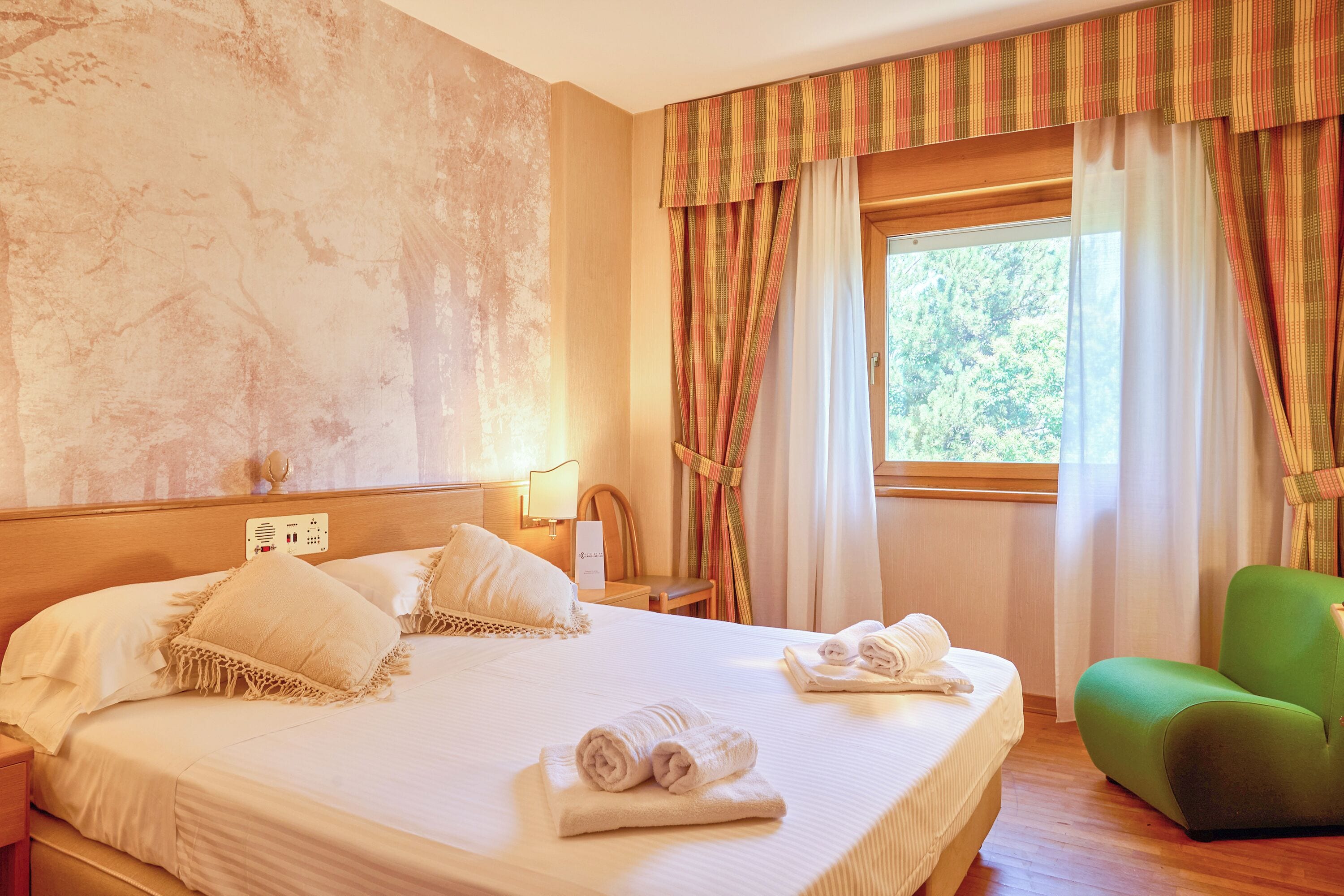 Habitació d'hotel amb un llit gran, un relaxant otomà verd i una finestra amb cortines.