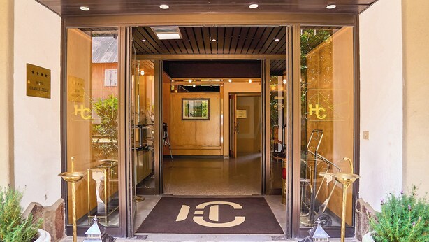 Ein Hotel Eingang mit Glastür, einer Metalldecke als Fußmatte und den Initialen des Hotels sowie einer kleinen Statue auf den Stufen.