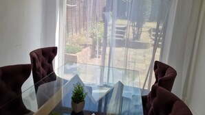 Ein Zimmer mit Glastisch und zwei dunklen Sesseln, einer kleinen Topfpflanze sowie Blick auf einen Innenhof mit Backsteinmauer und Grünpflanzen.