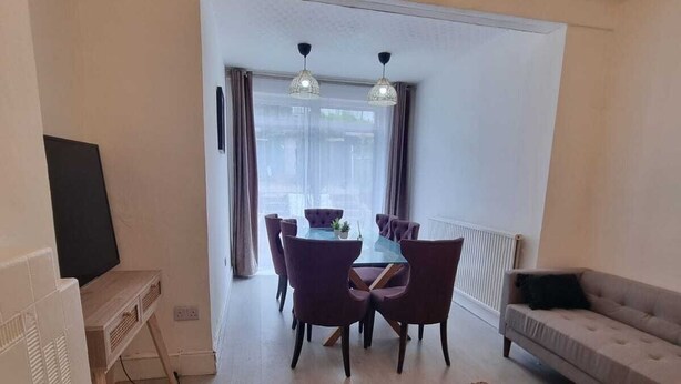 Ein Wohnzimmer mit einer Couch, einem Esstisch mit Stühlen, einem Fernseher und einem großen Fenster mit Vorhängen.