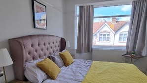 Ein Schlafzimmer mit Bett, Nachttisch, Lampe, einem Fenster mit Vorhängen und Blick auf Gebäude.