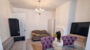 Ein modernes Wohnzimmer mit einem Glastisch, zwei violetten Sesseln, einem beigen Sofa, einem grauen Sessel, einem Fernseher und einer Kristallleuchter.