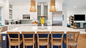 A modern kitchen with white cabinets, a large island with bar stools, a central island with a vase of flowers, and a stainless steel refrigerator. - Beach Access Directly Across Street! Ocean view, Private Pool, Hot Tub, Luxe New Construction (Isle Of Palms)