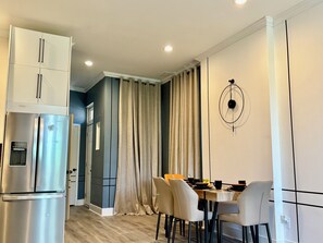 Une cuisine moderne dotée d’un réfrigérateur encastré, d’un coin repas avec une table et des chaises, et d’une grande fenêtre agrémentée de rideaux.