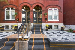 A building with a red brick facade, black and yellow striped steps leading to a set of double doors, and flower pots on the steps.