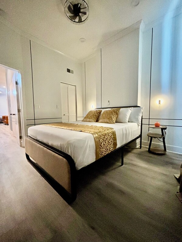 Une chambre à coucher moderne avec un grand lit, une table de chevet, un petit tabouret et un ventilateur de plafond.