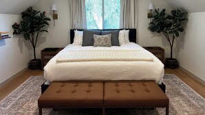 A bedroom with a large bed, two bedside tables, a bench, and two potted plants.