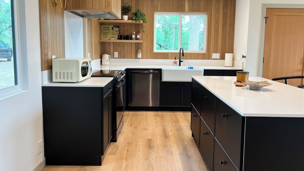 A modern kitchen with black cabinets, a white countertop, stainless steel appliances, and a window with a view of trees.