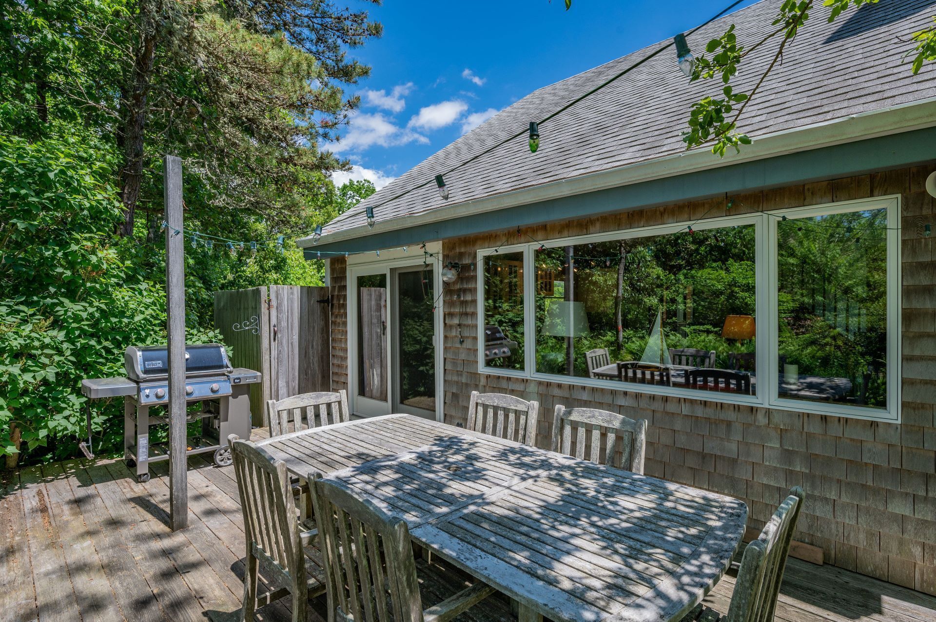 A covered patio with a wooden table and chairs, a grill, and a view of the surrounding greenery.