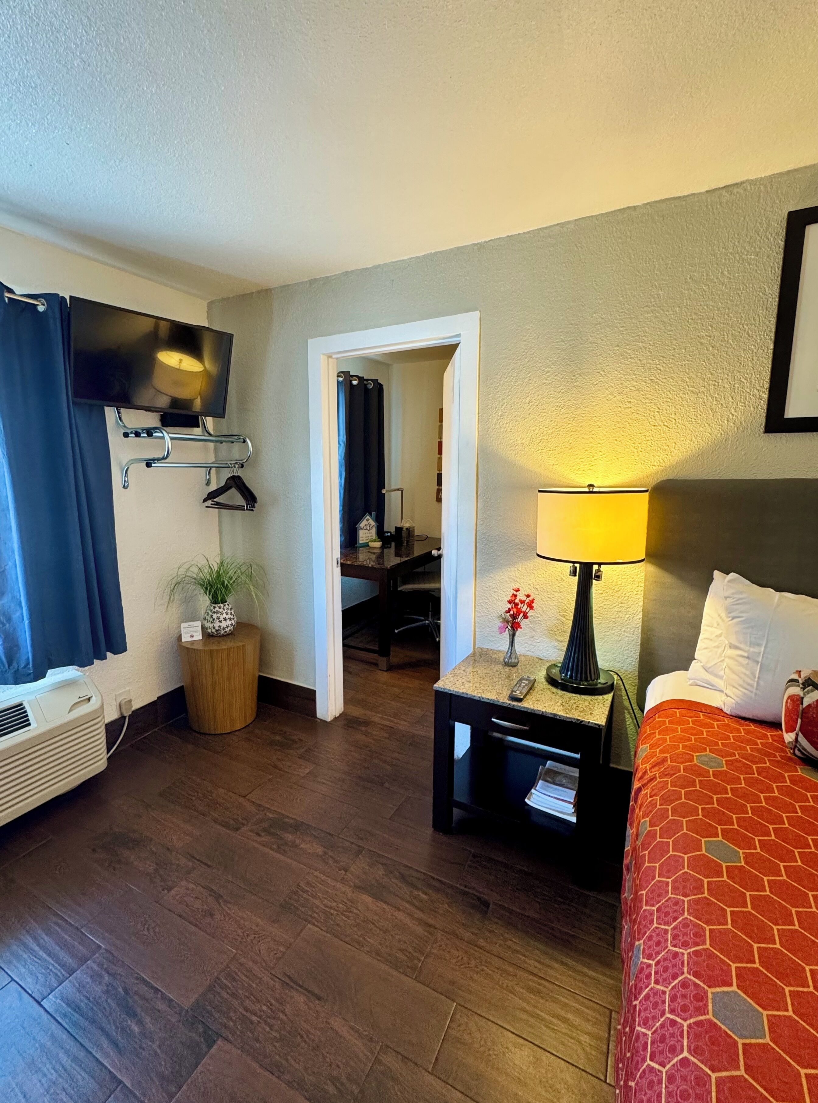 A hotel room with a bed, a TV mounted on the wall, a desk with a lamp, and a potted plant.