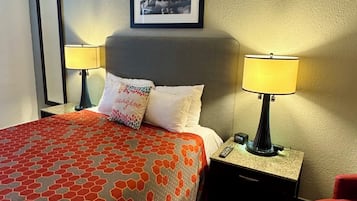 Una habitación de hotel con una cama, dos lámparas de noche, una mesita de noche, una silla, un cuadro en la pared y un espejo.