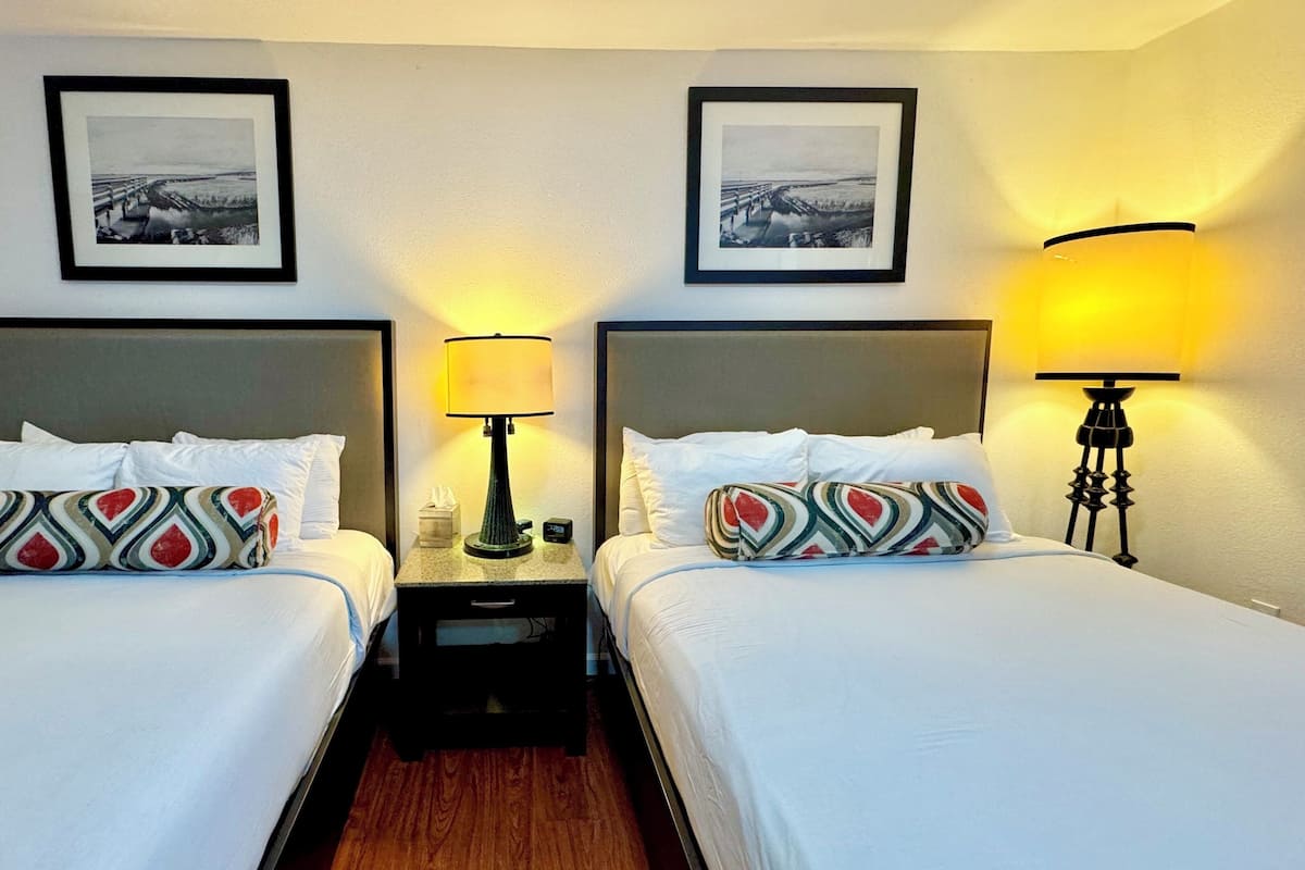 Une chambre d’hôtel avec deux lits, chacun agrémenté de draps blancs et d’oreillers à motifs, une table de chevet avec une lampe et des tableaux encadrés accrochés au mur.