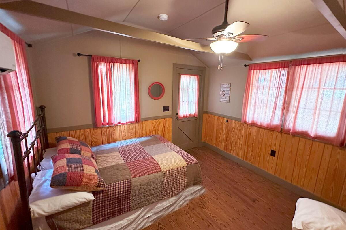 A bedroom with a wooden floor, a bed with a quilt, a ceiling fan, and pink curtains.