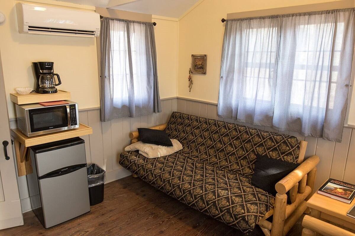 A compact living space with a kitchenette, including a microwave, coffee maker, and refrigerator. There is a patterned sofa with cushions and a small wooden table with a book.