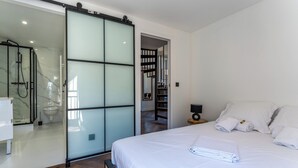 A bedroom with a bed, white linens, a glass shower enclosure, a wooden floor, and a hallway leading to a bathroom. - Duplex Bénézet - Welkeys (Avignon)