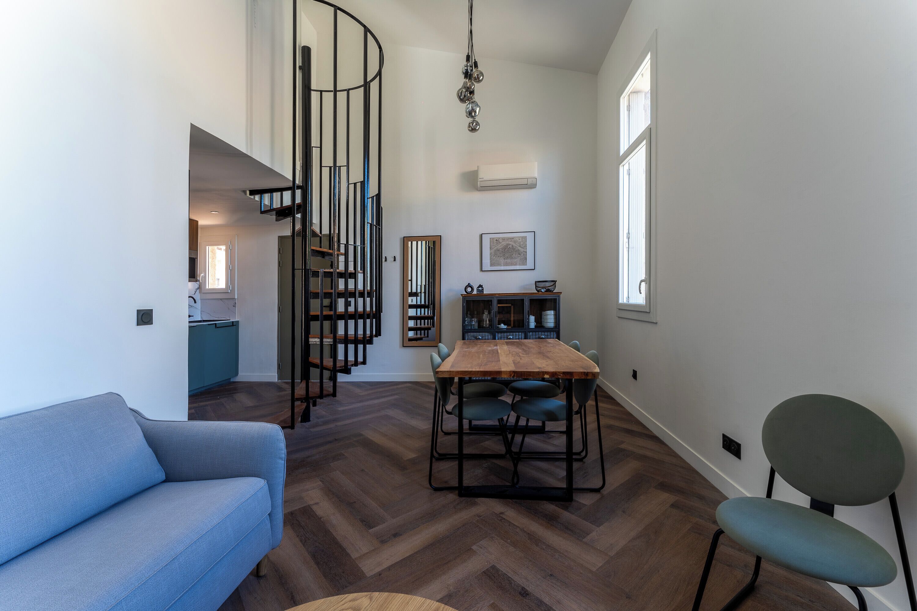 A modern living room with a blue sofa, a wooden dining table with chairs, a spiral staircase, and a view of a kitchen and dining area.