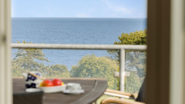A balcony with a view of the sea, a table with a fruit bowl, and a chair.