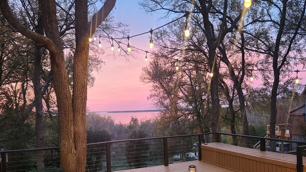 Eine Holzterrasse mit zwei Liegestühlen und einem Tisch, mit Blick auf ein Gewässer, im Hintergrund Bäume.