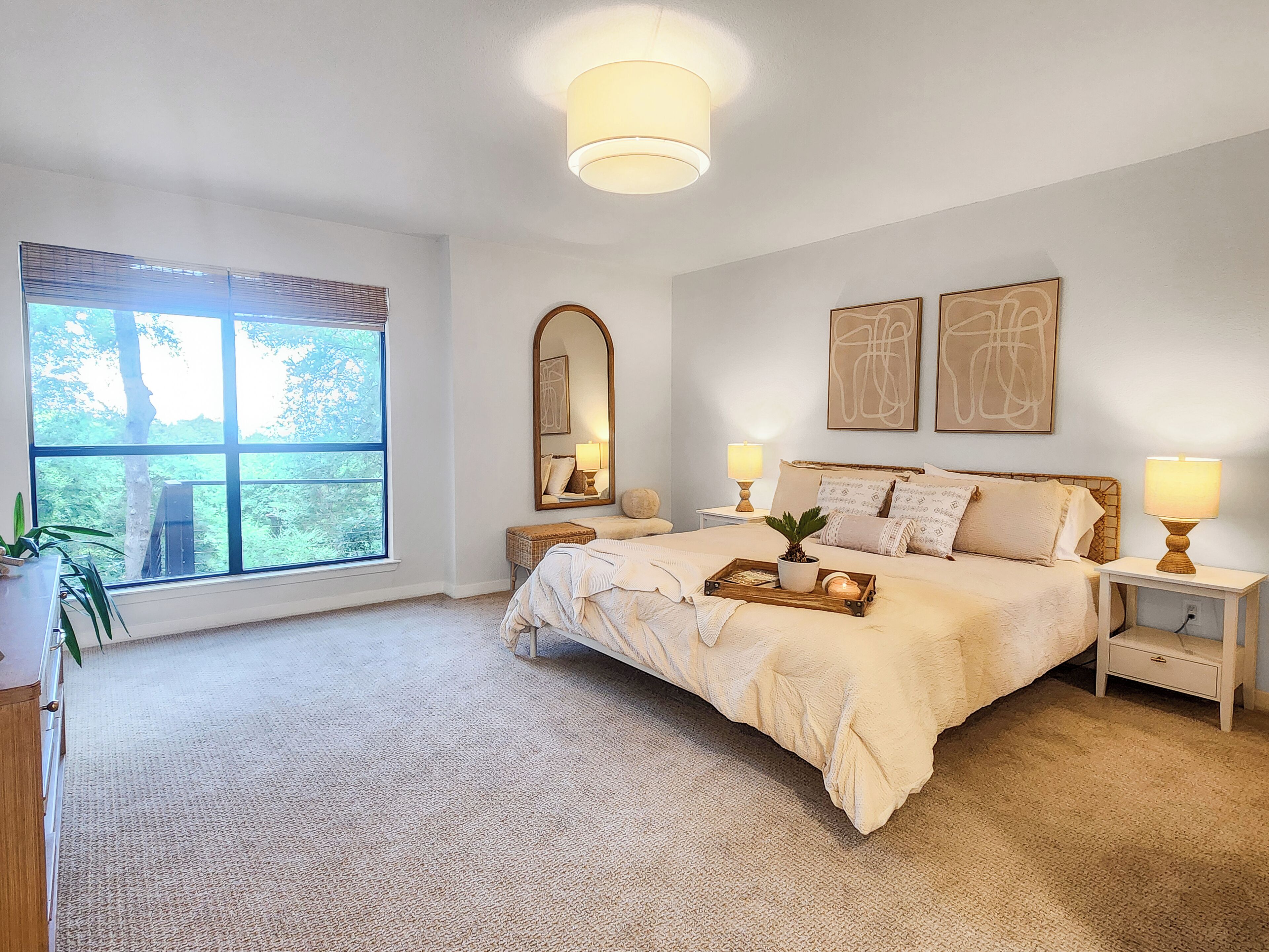 A bedroom with a large window, a bed with a tray of breakfast items, two bedside lamps, and a mirror on the wall.