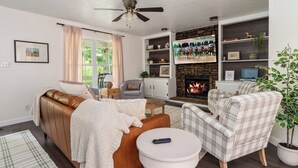 Una sala de estar con sofá, sillón y chimenea. Hay un televisor montado en la pared y un ventilador de techo.