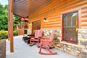 A log cabin with a porch, stone wall, and two rocking chairs. - Hidden Vista Lodge | Hot Tub | Game Room | Views! (Black Mountain)