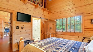 Ein Schlafzimmer in einer Blockhütte mit einem Bett, einem Fernseher, einem Fenster und einer Tür zu einem anderen Raum.