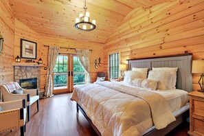 Een slaapkamer in een houten hutje, met een bed, een stoel, een open haard en een raam met gordijnen.