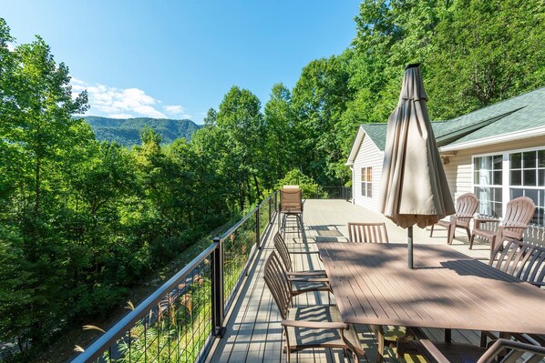A deck with outdoor furniture, a table, and chairs, overlooking a forested area with a house in the background.
