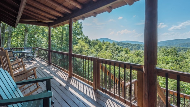 A wooden deck with outdoor furniture, overlooking a forested landscape with distant hills.