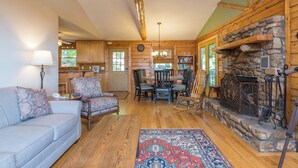 A cozy cabin interior with a stone fireplace, wooden walls, a dining area, and a living room with a sofa and patterned cushions.