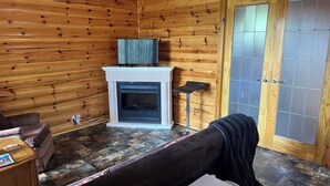 Interior de una cabaña de troncos con chimenea, un sofá, un televisor y una puerta con panel de vidrio esmerilado.