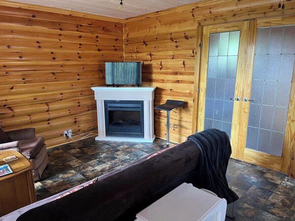Interior de una cabaña de troncos con chimenea, un sofá, un televisor y una puerta con panel de vidrio esmerilado.