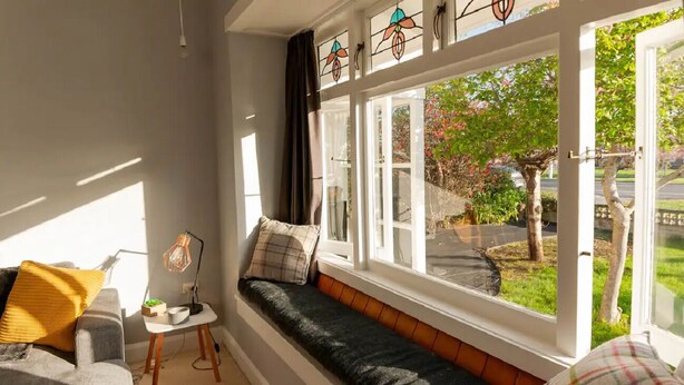Een gezellige kamer met een inbouwkruk, een klein tafeltje met een lamp, en een uitzicht op een tuin door grote ramen.