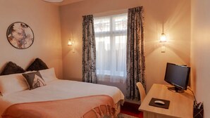 Een slaapkamer met een bed, een bureau met een computer, een televisie en een raam met gordijnen.