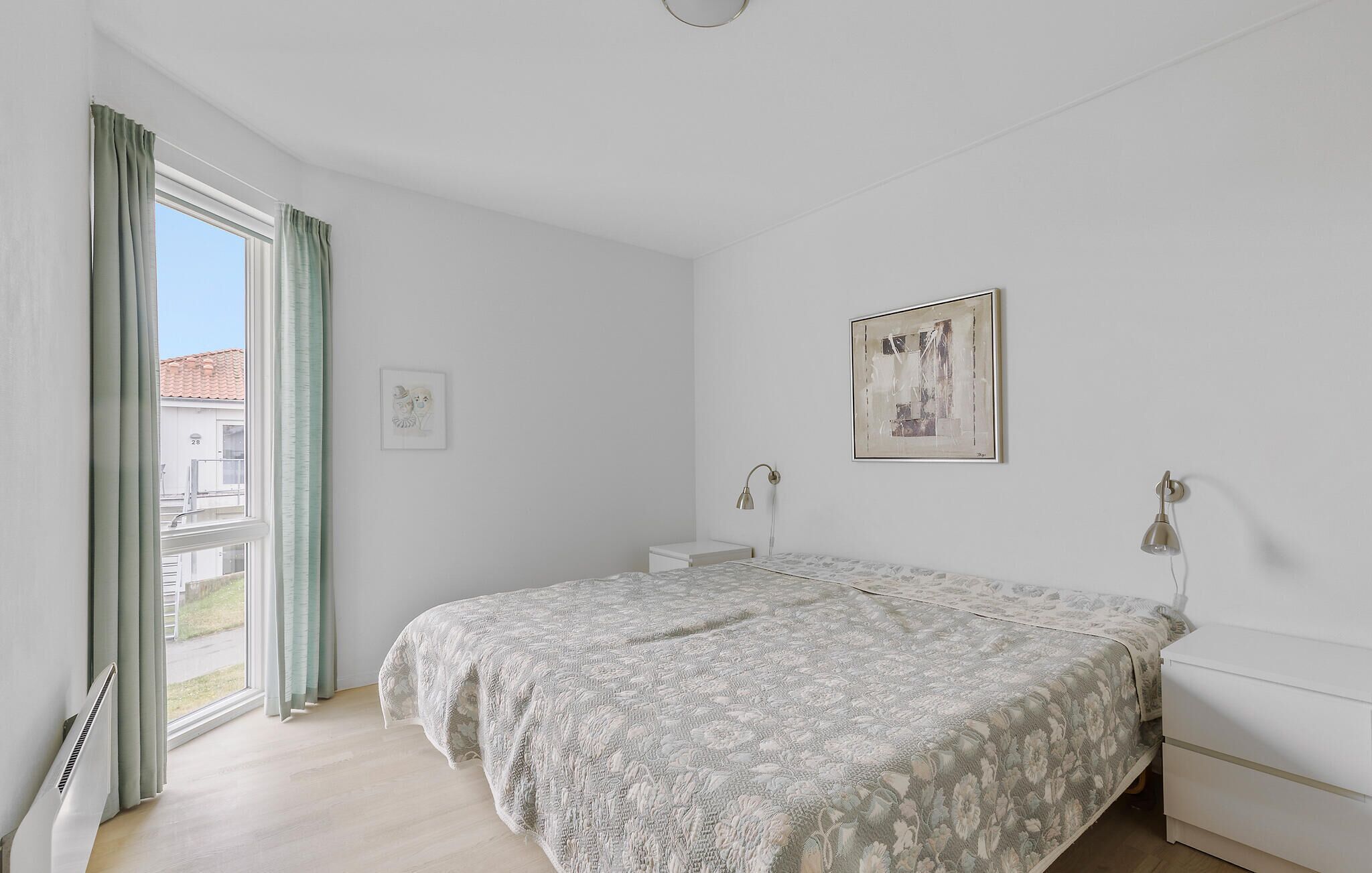 Une chambre à coucher avec un lit, une table de chevet, une lampe, une fenêtre avec des rideaux et un tableau encadré au mur.