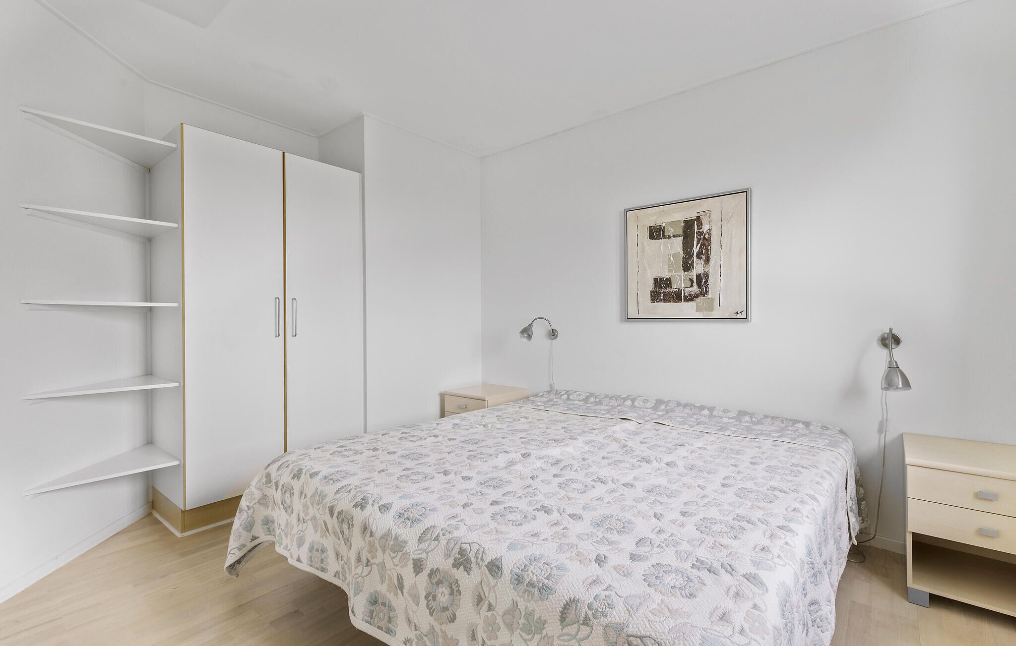 A bedroom with a bed, a wardrobe, a nightstand, a desk, and a wall-mounted light fixture.